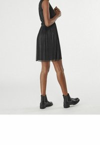 Mustapuku, jossa on hihaton malli, istuva vyötärö ja laskostettu hame, yhdistetty mustien nilkkasaappaiden kanssa, joissa on hopeiset yksityiskohdat.