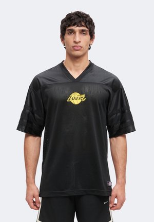 Młody mężczyzna ubrany w czarny siatkowany trykot Los Angeles Lakers i czarne spodenki Nike, stojący na tle jednolitego jasnego tła.