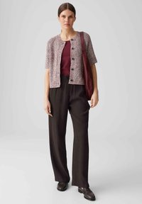 Strickjacke in Mauve mit Knopfverschluss, kombiniert mit einem bordeauxfarbenen Oberteil und schwarzen, gerippten, weiten Hosen. Die Accessoires umfassen eine burgunderfarbene Tasche.