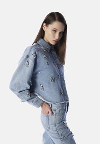 Giacca di jeans con decorazioni argentate, maniche lunghe, colletto e orlo sfrangiato, abbinata a pantaloni di jeans coordinati.