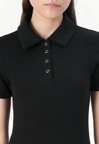 Black ribbed polo shirt featuring a collared neckline with six metal snap buttons, short sleeves, and a fitted silhouette.