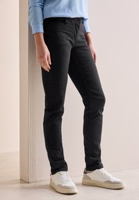 Jeans de mezclilla negros con un ajuste slim, que cuentan con bolsillos frontales y una textura suave, combinados con zapatillas deportivas blancas y una camiseta azul clara.