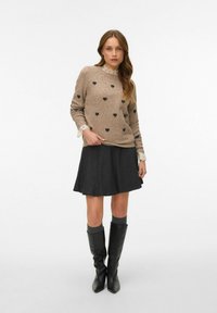 Beiger Pullover mit schwarzen Herzmustern, Spitzenbündchen; kombiniert mit einem dunklen Rock und knielangen schwarzen Lederstiefeln. Einfache Texturen und Formen.