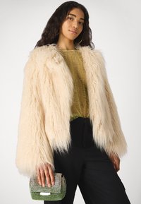 Chaqueta de pelo sintético en crema con mangas largas y texturizadas; combinada con una blusa verde transparente y pantalones negros. Sosteniendo un bolso de mano con degradado brillante.
