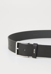 Cintura in pelle nera con finitura texture, fibbia argentata e dettaglio del logo. Presenta diversi fori per la regolazione della misura.