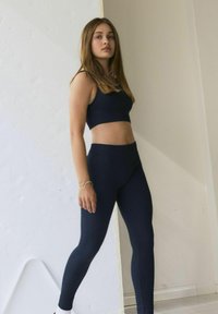 Jūros mėlynos sportinės liemenėlės ir aukštu juosmeniu leggingsai iš lygaus, tampraus medžiagos. Modelis stovi pasitikėdamas savimi neutralioje fone.