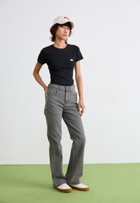 Camiseta negra ajustada, jeans gris de pierna recta y gorra beige con logo rojo. Zapatillas blancas con suelas marrones claras sobre un suelo verde.