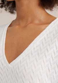 Top bianco con scollatura a V che presenta un motivo a spina di pesce testurizzato, con dettagli di cucitura discreti e un tessuto leggero e traspirante.