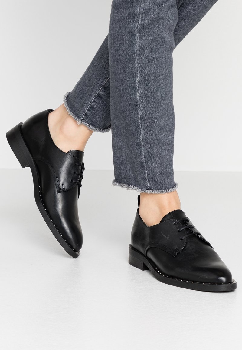 Chaussures à lacets en cuir noir portées avec un jean gris à ourlet effiloché sur un sol blanc, montrant les deux pieds de la mi-mollet vers le bas.