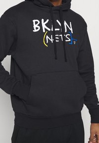 Svart huvtröja med en framficka, prydd med vit, blå och gul text "BKLYN (NETS)" tvärs över bröstet och justerbara svarta snören.