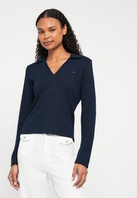 Marineblå langermet polo med V-hals og krage. Har en liten logo på brystet og er kombinert med hvite bukser. Glatt tekstur.