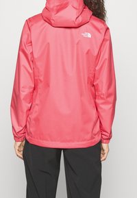 Chaqueta impermeable rosa con capucha, puños ajustables y logo en la parte trasera, con una textura lisa y brillante y un corte holgado.