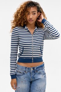 Jonge vrouw met krullend haar in een blauw-wit gestreepte ritstrui en versleten blauwe spijkerbroek, die poseert met één hand bij haar oor.