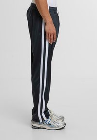 Fekete sportnadrág, két függőleges fehér csíkkal. Simább, könnyű anyagból készült. Rugalmas derékkal és oldalsó zsebekkel rendelkezik.