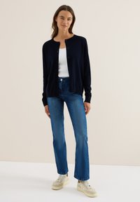 Marineblauwe cardigan van zachte stof, met een ronde halslijn, knopen aan de voorkant en ribgebreide manchetten; gecombineerd met blauwe uitlopende jeans en beige sneakers.