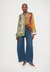 Kolorowa jedwabna koszula z wzorem paisley, szerokimi rękawami i kołnierzem, zestawiona z niebieskimi, szerokimi spodniami z wysokim stanem.