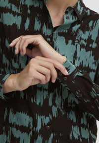 Svart button-up skjorta med turkos abstrakt mönster. Långärmad med knäppning vid ärmslut. Slät tyg, strukturerad krage och synliga knappar.