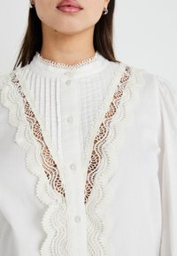 Blouse blanche avec un tissu plissé et une bordure en dentelle le long de l'encolure et du devant, fermeture par boutons, et manches légèrement bouffantes.