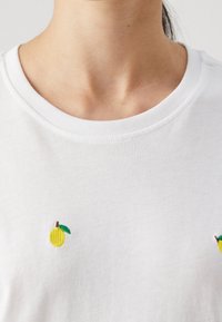 Bílá bavlněná tričko s malou vyšívanou žlutou citronem a zelenými listovými akcenty na hrudi. Kulatý výstřih, měkká textura.