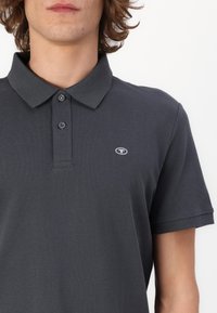 Polo shirt gri închis, cu guler clasic, placket cu două butoane și un mic logo pe partea stângă a pieptului. Material texturat și mâneci scurte.