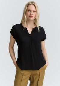 Schwarze kurzärmlige Bluse mit V-Ausschnitt und einer Falte vorne, aus glattem Stoff, kombiniert mit hellbraunen Hosen.