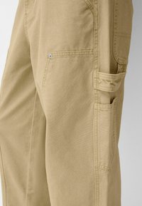 Pantalones cargo color caqui hechos de sarga de algodón, con múltiples bolsillos, una textura suave y costuras reforzadas a lo largo de las uniones.