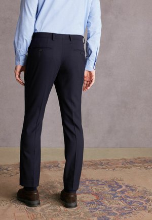 Tmavomodré nohavice so slim fit strihom, s dvoma zadnými vreckami na gombíky a čistým, prispôsobeným dizajnom, skombinované s hnedými topánkami.