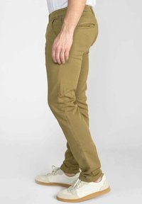 Man in slim-fit kaki broek en witte sneakers, zijwaarts staand tegen een effen lichte achtergrond.