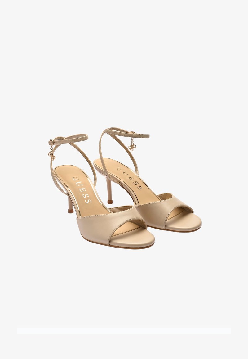 Sandales à talons hauts beiges à bout ouvert avec brides à la cheville et détails de charms dorés, logo de la marque GUESS sur les semelles intérieures.