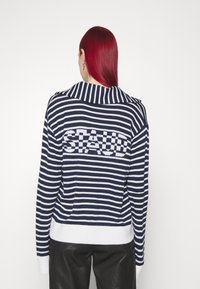 Marineblauwe en witte gestreepte gebreide trui met een brede kraag, geribde manchetten en het "STAUD"-logo in blokletters op de achterkant. Glad textuur.
