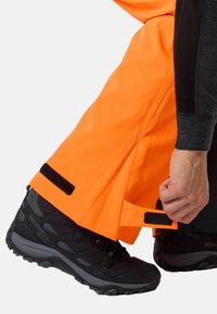 Oranje waterdichte broek van duurzaam materiaal, met verstelbare klittenbandriemen bij de zoom en gecombineerd met zwarte wandelschoenen.