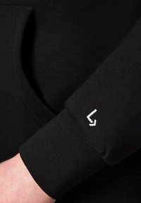 Czarny sweatshirt o miękkiej teksturze, z mankietem z białym haftowanym logo. Widoczna kieszeń kangurka, podkreślająca prostotę designu.