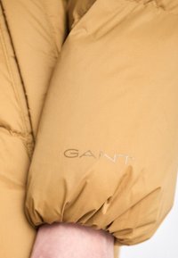 Kreminės spalvos pūkuota striukė su švelniu apdaru, turinti elastinius rankogalius ir sidabrinę "GANT" logotipą ant rankovės.