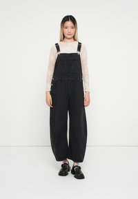 Črne denim naramenice širokih nogavic z nastavljivimi naramnicami, v kombinaciji z lahnim čipkastim dolgim rokavom in črnimi čevlji.