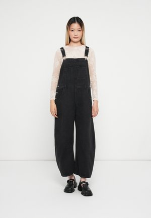 Svarta denimsoveraller med vida ben och justerbara axelremmar, matchade med en ljus spetsblus med långa ärmar och svarta skor.