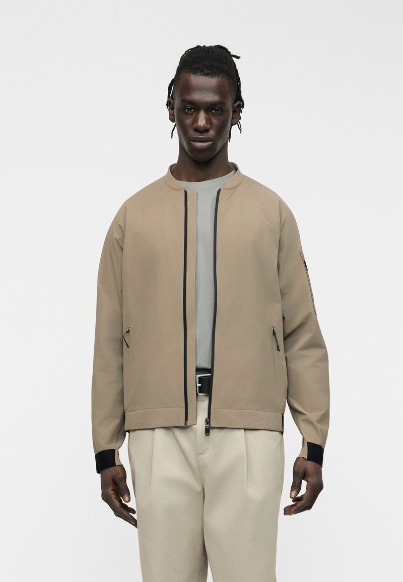 Jeune homme portant une veste bomber beige sur une chemise grise et un pantalon beige clair, debout devant un fond blanc uni.