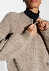 Beige, fuzzy gebreide cardigan met een lichte glans, voorzien van een hoge kraag en aansluitende mouwen. De draagster sluit het knoopje, waardoor de textuur zichtbaar is.