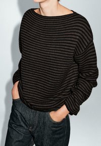 Osoba w luźnym, długim rękawie, czarno-brązowym swetrze w paski, włożonym w ciemnoniebieskie dżinsy, z rękami w kieszeniach.