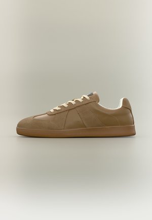 Brauner Sneaker mit niedrigem Schaft aus Wildleder- und Lederpaneelen, cremefarbenen Schnürsenkeln und flacher Gummisohle, seitlich auf neutralem Hintergrund gezeigt.
