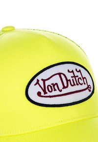 Gorra de béisbol amarilla con un parche ovalado blanco que lleva el texto "Von Dutch" en rojo. El material parece suave y tiene un diseño estructurado.