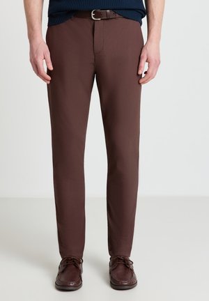 Mann trägt braune Slim-Fit-Hose, passenden braunen Gürtel und braune lederne Schnürschuhe, steht vor einfachem hellem Hintergrund.
