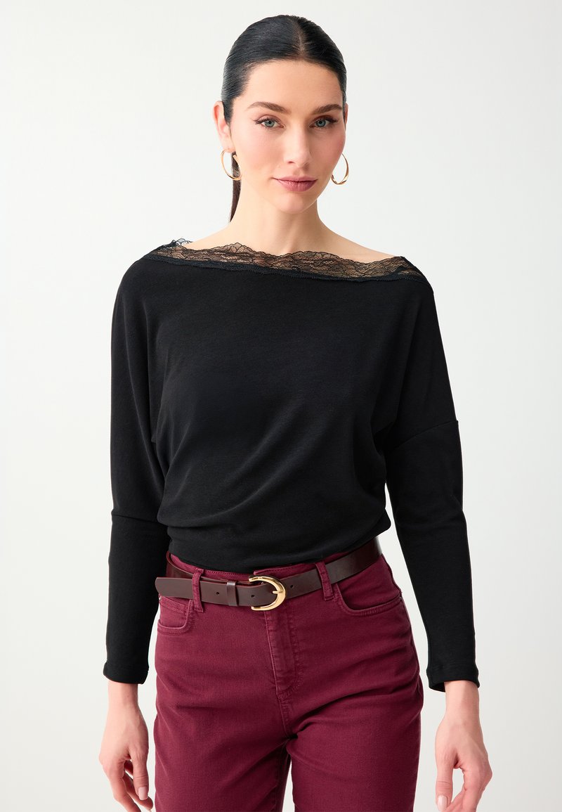 Femme portant un haut noir à manches longues avec un col en dentelle, un pantalon bordeaux, des boucles d'oreilles créoles dorées et une ceinture rouge foncé avec une boucle dorée.