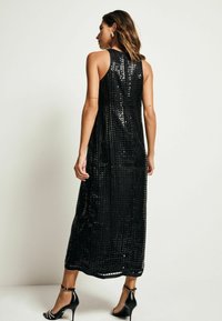 Robe maxi noire pailletée avec un design sans manches. Le tissu présente un motif texturé et elle est associée à des sandales noires à talons hauts.