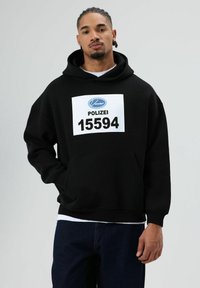 Zwarte capuchontrui met buidelzak aan de voorkant, met een groot wit label met de tekst "POLIZEI 15594" en een logo. Zachte textuur, losse pasvorm.