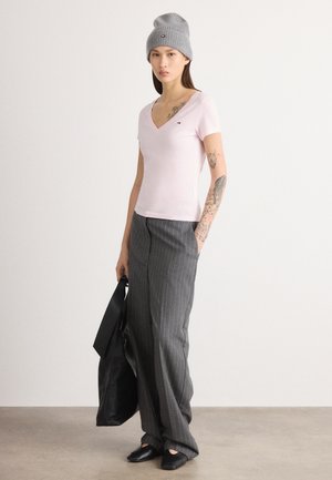 Rosa v-hals t-skjorte, grå beanie, stripete bukser og svarte sko. Modellen holder en svart totebag. Tatoveringer er synlige på armen.