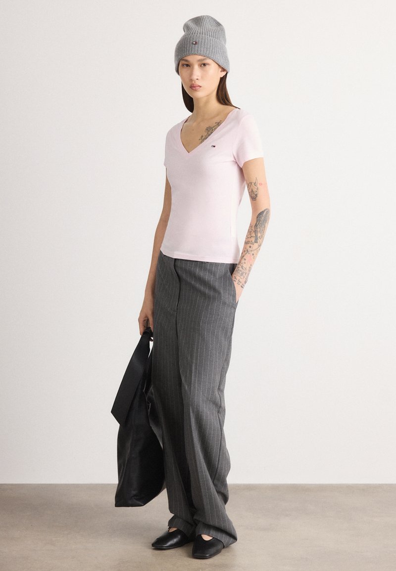 Ροζ μπλουζάκι με V-λαιμό, γκρι σκουφάκι, παντελόνι με ρίγες και μαύρα παπούτσια. Το μοντέλο κρατάει μια μαύρη τσάντα ώμου. Τατουάζ είναι ορατά στο χέρι.