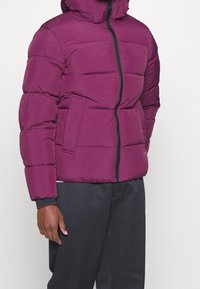 Fioletowa kurtka puchowa z kapturem, pikowanym wzorem i suwakiem z przodu. Posiada boczne kieszenie i ściągacze przy rękawach, wykonana z lekkiego materiału.