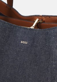 Denim-kassi ruskeilla nahkaosilla, jossa on vetoketjukiinnitys ja kultainen "BOSS"-logo. Rakenteellinen kangas, jossa näkyvät ompelukuviot.