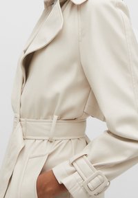 Trench-coat beige avec une ceinture à la taille, un large col à revers et des manches ajustées ornées de boucles. Texture de tissu lisse.