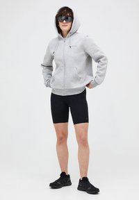 Grauer Reißverschluss-Hoodie mit einer Kapuze zum Zusammenziehen und Fronttaschen, kombiniert mit schwarzen Radshorts und schwarzen Sportschuhen. Dunkle Sonnenbrille getragen.